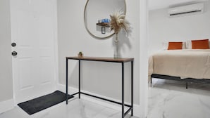 A room with a white door, a wooden console table with a mirror, a small shelf with decorative items, and a bed with orange pillows.