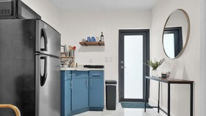 A compact kitchen with a black refrigerator, blue cabinets, a small shelf with kitchenware, a trash bin, and a mirror on the wall.
