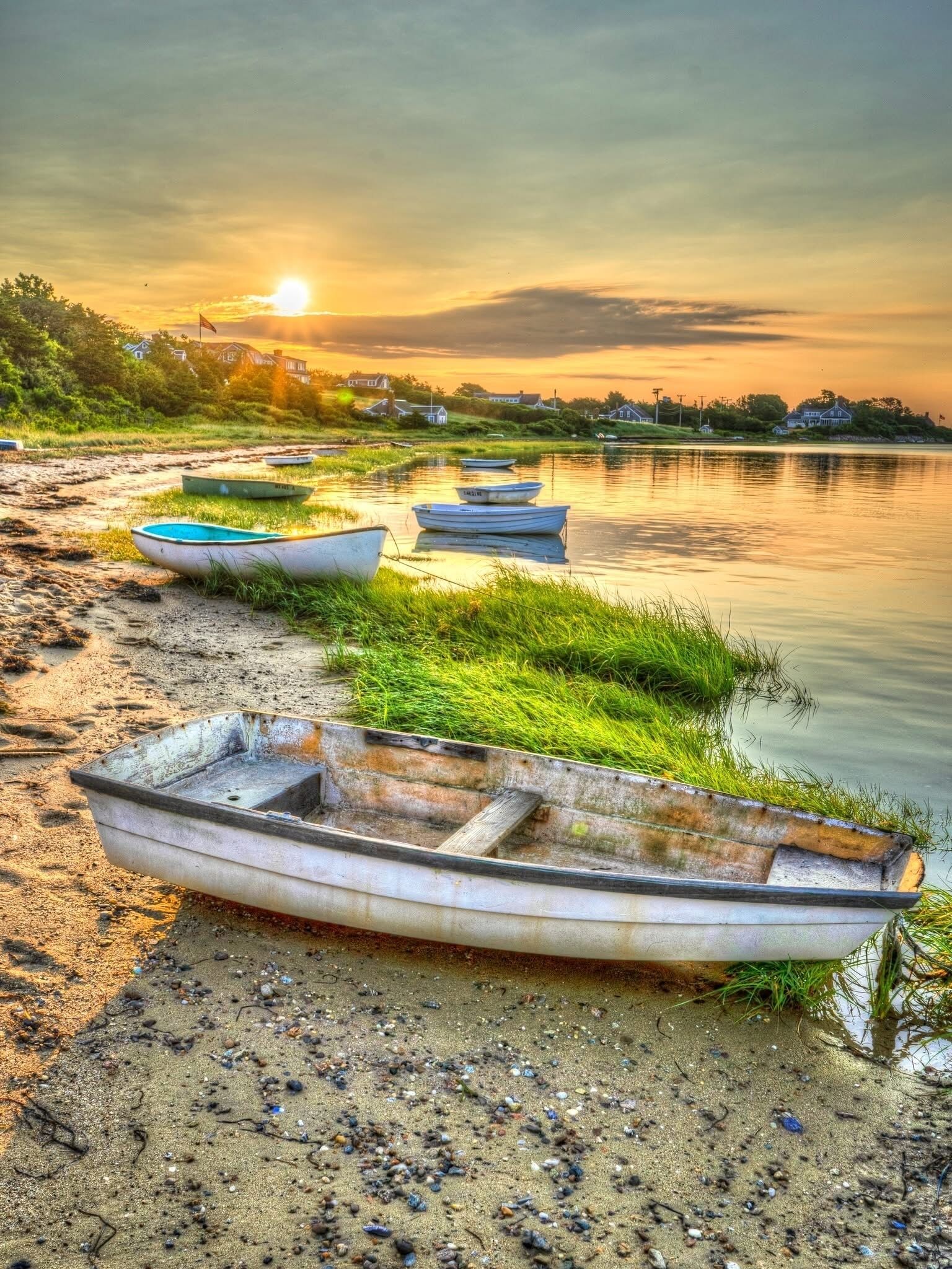 A row of small boats lined up on a sandy shore next to a calm body of water during sunset.