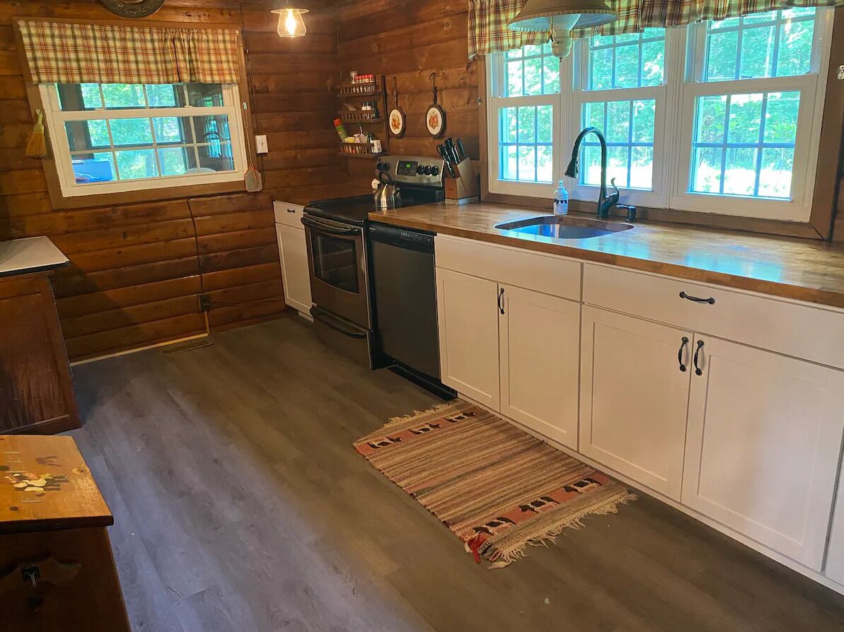 Une cuisine de style rustique, avec des éléments en bois, comprenant un lave-vaisselle, une cuisinière et une grande fenêtre agrémentée d’une lambrequin à carreaux.