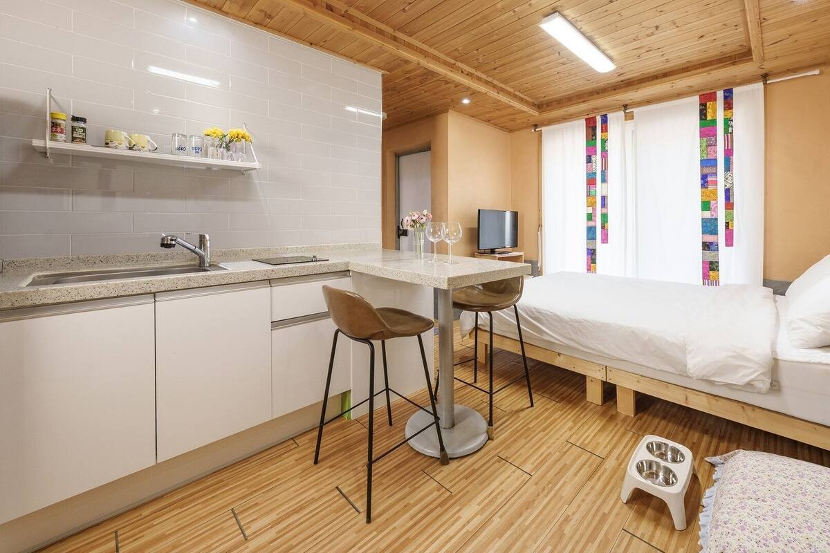 Een kleine kamer met een kitchenette, een bed, een televisie en een raam met kleurrijke gordijnen.