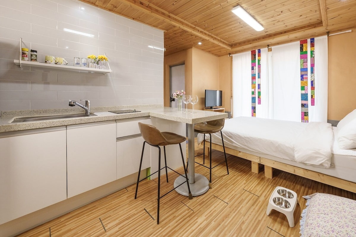 A compact room with a kitchenette, a bed, a TV, and a window with colorful curtains.