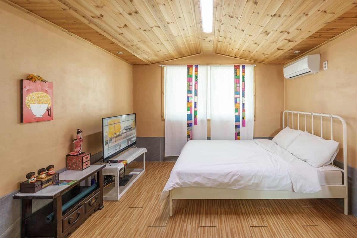 Een slaapkamer met een houten plafond, een bed, een televisie, een dressoir en een raam met gordijnen.