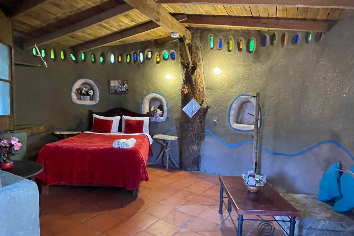 A rustic room with a wooden ceiling, a bed with red bedding, a small table, and a sofa with blue cushions.