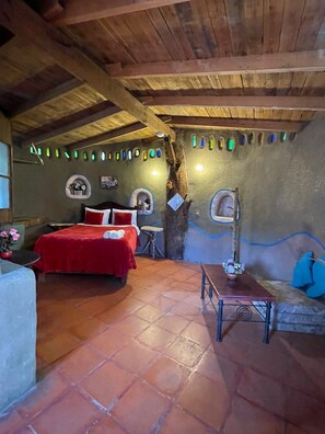 A rustic room with a wooden ceiling, a bed with red bedding, a small table, and a sofa with blue cushions.