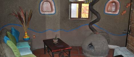 A rustic room with a stone wall, a wooden table, a sofa with colorful cushions, a small window, and a stone oven.