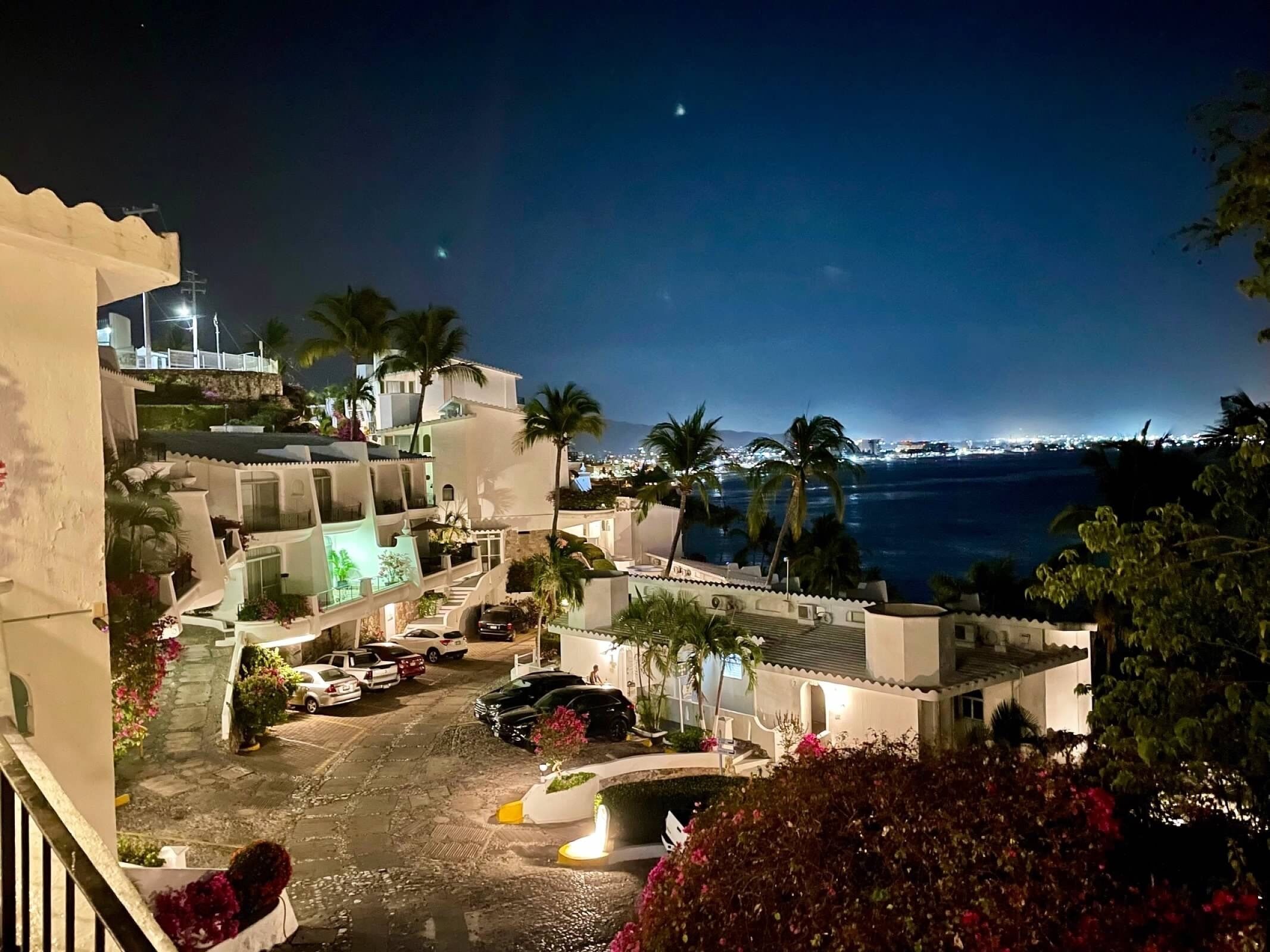 A coastal hotel with multiple balconies, a parking area, and a view of the city and ocean at night.