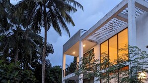 A modern two-story house with a balcony, surrounded by lush greenery and palm trees.