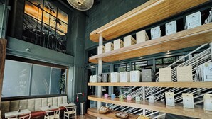 Una cafetería con estantes de madera que exhiben sacos de café, una zona de estar con bancos acolchados y un amplio ventanal con vistas a palmeras.