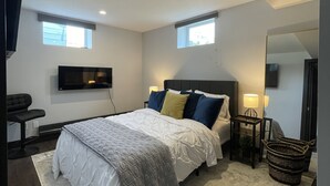A modern bedroom with a large bed, a TV mounted on the wall, a small desk, and a window. - The Lux Den Speaks for itself. Cozy, bright self-contained basement suite  (Yorkton)