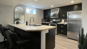 A modern kitchen with a central island, black cabinets, stainless steel appliances, and a wooden floor. - The Lux Den Speaks for itself. Cozy, bright self-contained basement suite  (Yorkton)