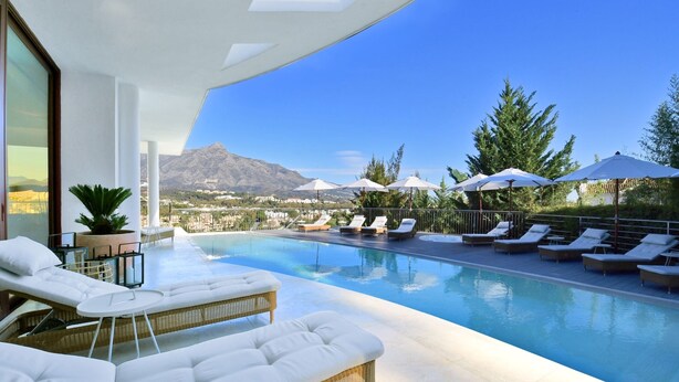 A modern outdoor pool area with lounge chairs and umbrellas, a clear view of mountains, and a poolside table. - Extraordinary 7 BDRM Villa w Pool, Gym, Cinema (Marbella)