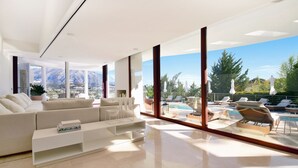 A modern living room with a sofa, coffee table, and large windows overlooking a pool and mountains. - Extraordinary 7 BDRM Villa w Pool, Gym, Cinema (Marbella)