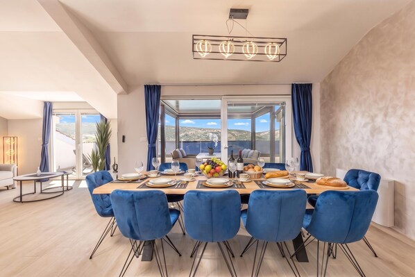A dining area with a large table set for six, blue upholstered chairs, and a view of the outdoors through large windows.
