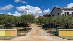 Un camino rural que lleva a una casa blanca con techo rojo, rodeada de vegetación y coches aparcados.