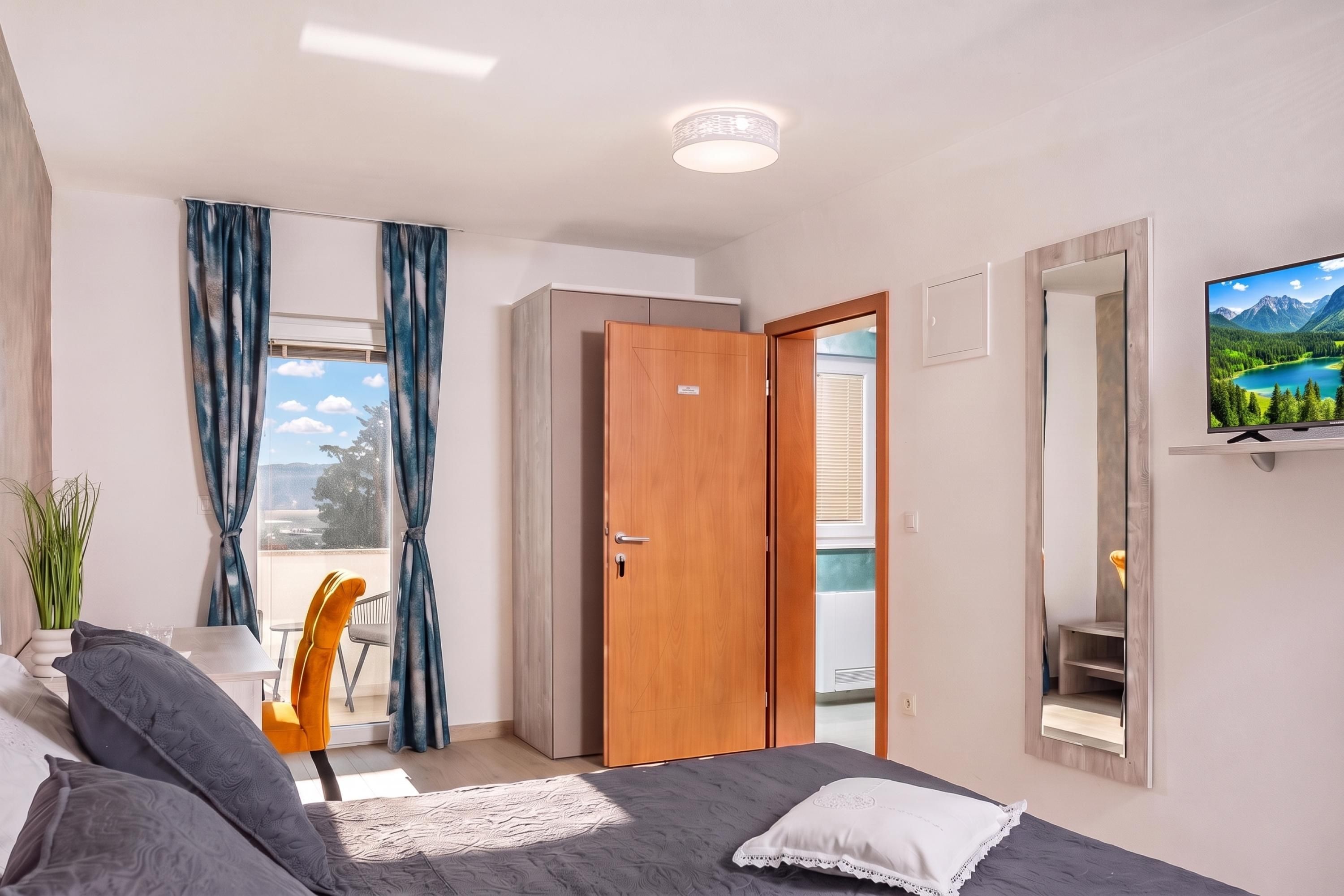 A modern bedroom with a bed, a TV, a balcony with a chair, and a view of the outdoors.
