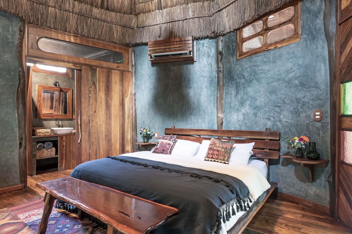 A rustic bedroom with a wooden bed, a small bedside table, a bench, and a thatched ceiling.