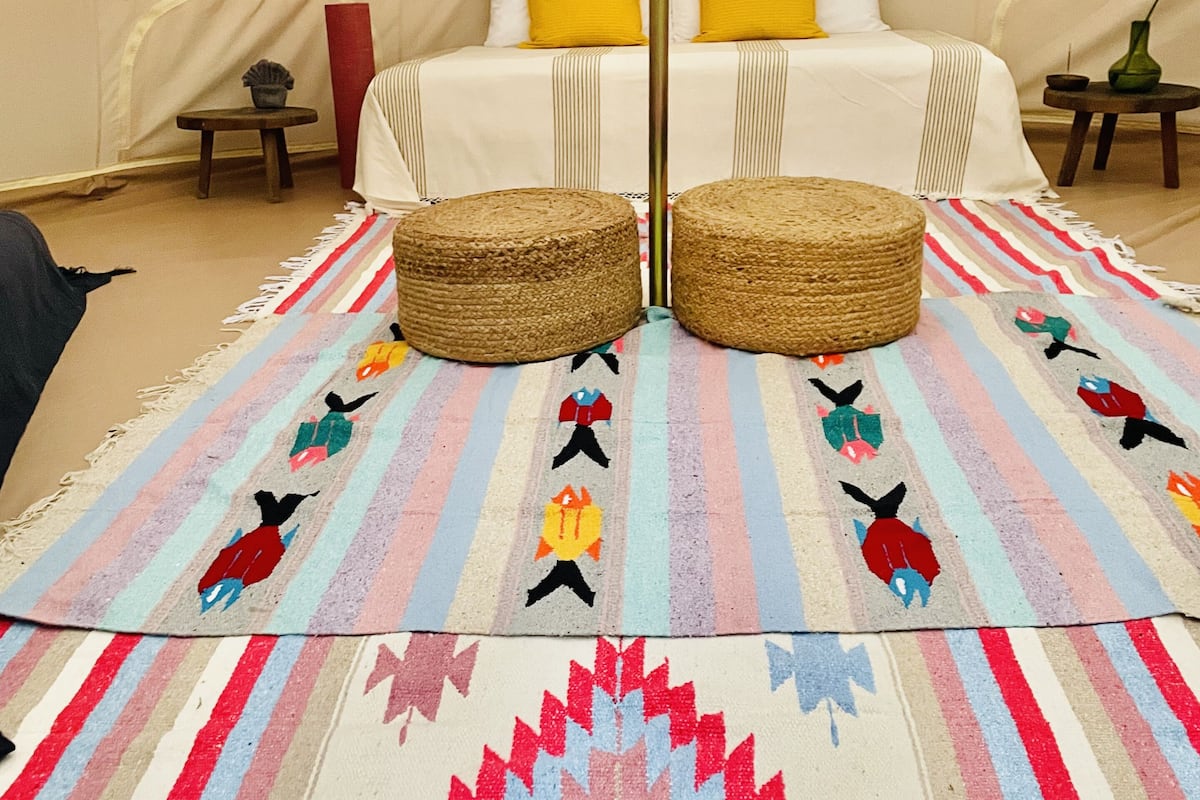 A tented accommodation with a bed, two woven ottomans, a colorful striped rug, and a small table with a vase.