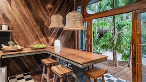 Una zona de bar con barra de madera, suelo a cuadros, dos lámparas de mimbre colgantes y vistas a plantas tropicales a través de amplios ventanales.