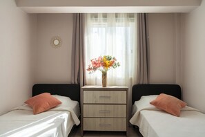 Une chambre avec deux lits, une table de chevet ornée d’un vase de fleurs et une fenêtre avec des rideaux légers.