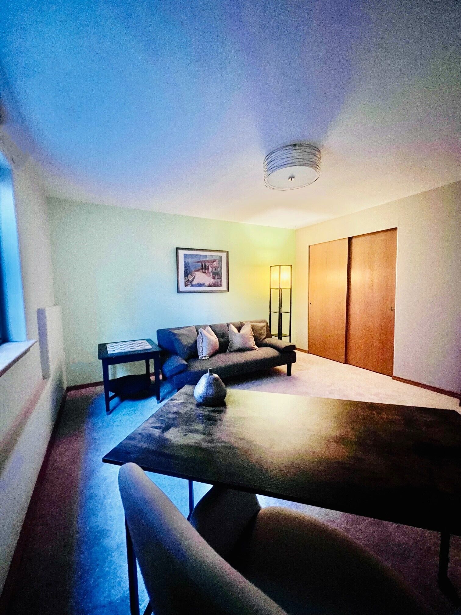 A modern living room with a sofa, a coffee table, and a dining table. There is a painting on the wall and a lamp on the side table.