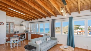 Un vaste espace de vie comprenant un canapé, une table à manger et des chaises. Il y a également un coin cuisine équipé d’un four à micro-ondes et de placards. De grandes fenêtres offrent une vue sur la mer et sur une colline.