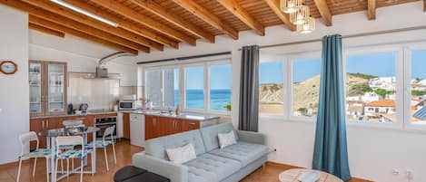 A spacious living area with a sofa, a dining table, and chairs. There is a kitchen area with a microwave and cabinets. Large windows provide a view of the sea and a hillside.
