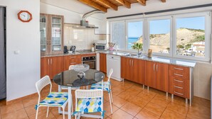 Une cuisine moderne avec des meubles en bois, une table à manger avec des chaises et une vue sur la mer grâce à de grandes fenêtres.