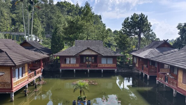 A traditional wooden house with a tiled roof, situated on stilts over a body of water, surrounded by lush greenery and trees.
