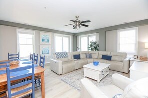 A spacious living room with a beige sectional sofa, a dining table with blue chairs, a ceiling fan, and large windows.