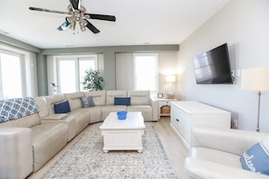 A living room with a beige sectional sofa, a white coffee table, a ceiling fan, a flat-screen TV mounted on the wall, and a large window providing natural light.