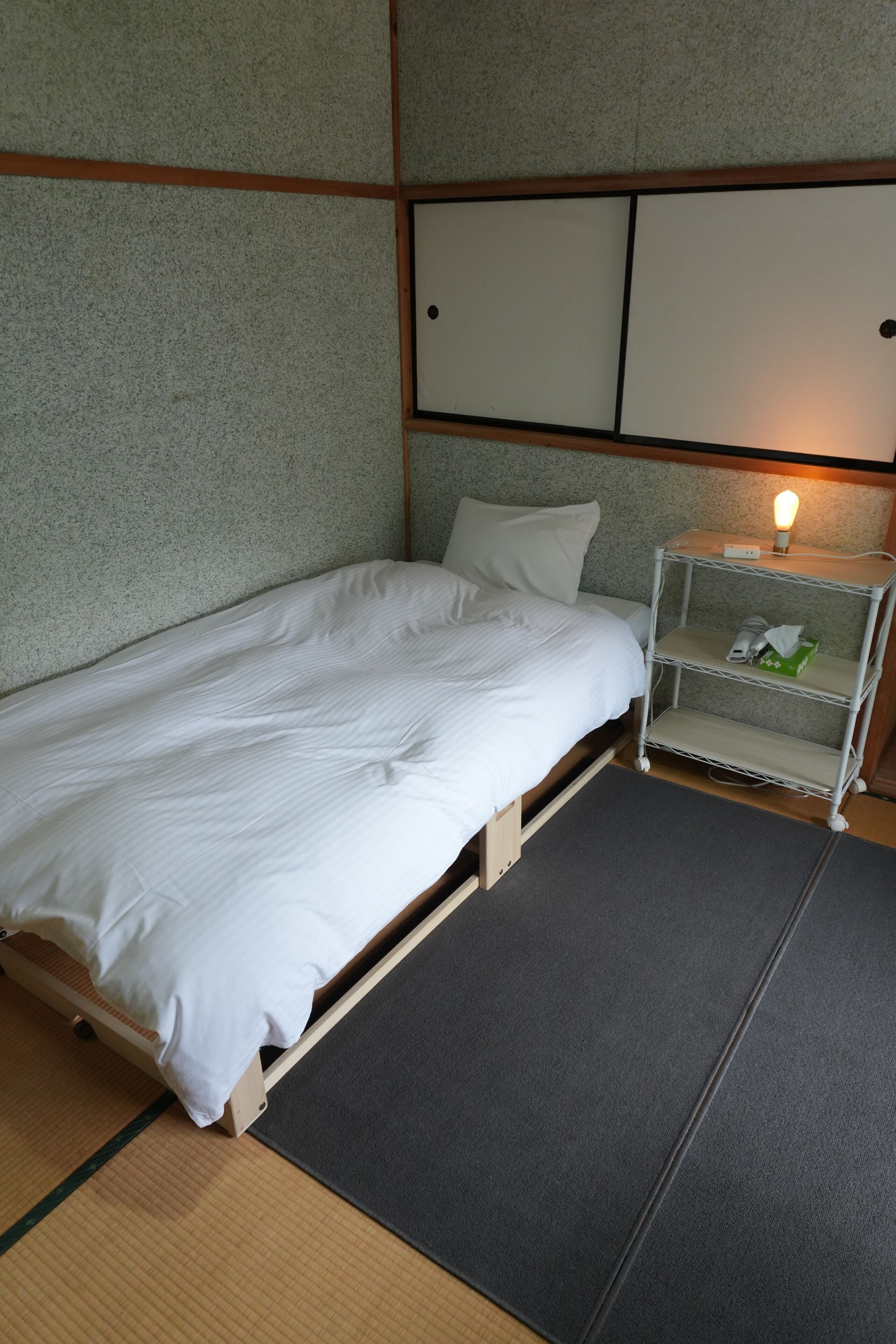 A single bed with a wooden frame and white bedding in a room with a textured wall and a small bedside table with a lamp.