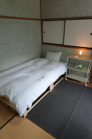 Una cama individual con estructura de madera y ropa de cama blanca en una habitación con pared texturizada y una mesita de noche pequeña con lámpara.