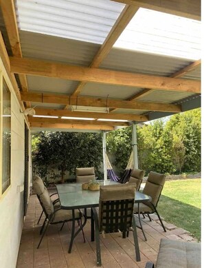 Une terrasse couverte avec une table et des chaises, des poutres en bois et une vue sur la cour.