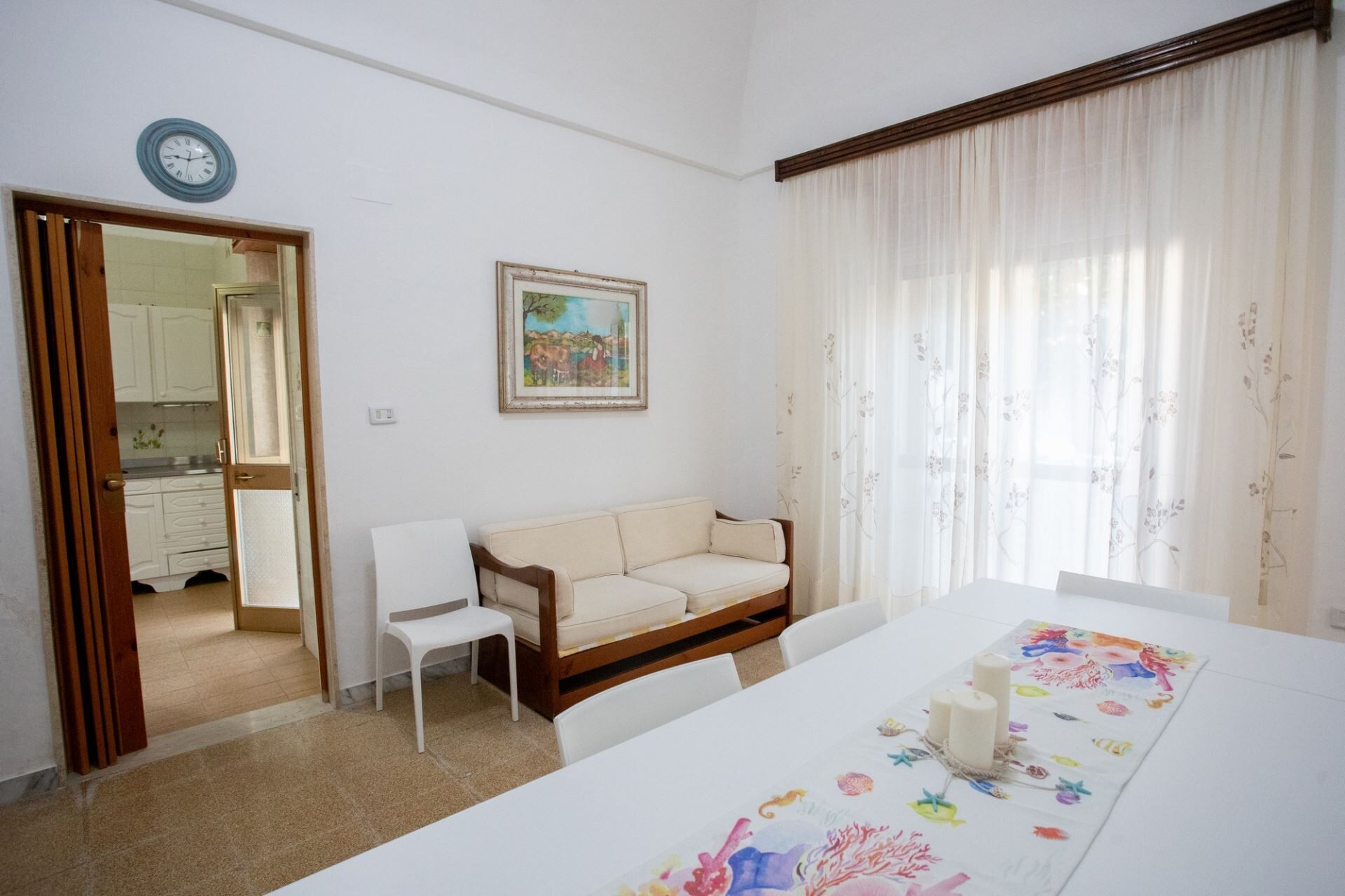 A room with a white sofa, a dining table with a floral table runner, a painting on the wall, and a window with sheer curtains.