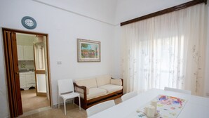 A room with a white sofa, a dining table with a floral table runner, a painting on the wall, and a window with sheer curtains.