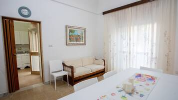 A room with a white sofa, a dining table with a floral table runner, a painting on the wall, and a window with sheer curtains.