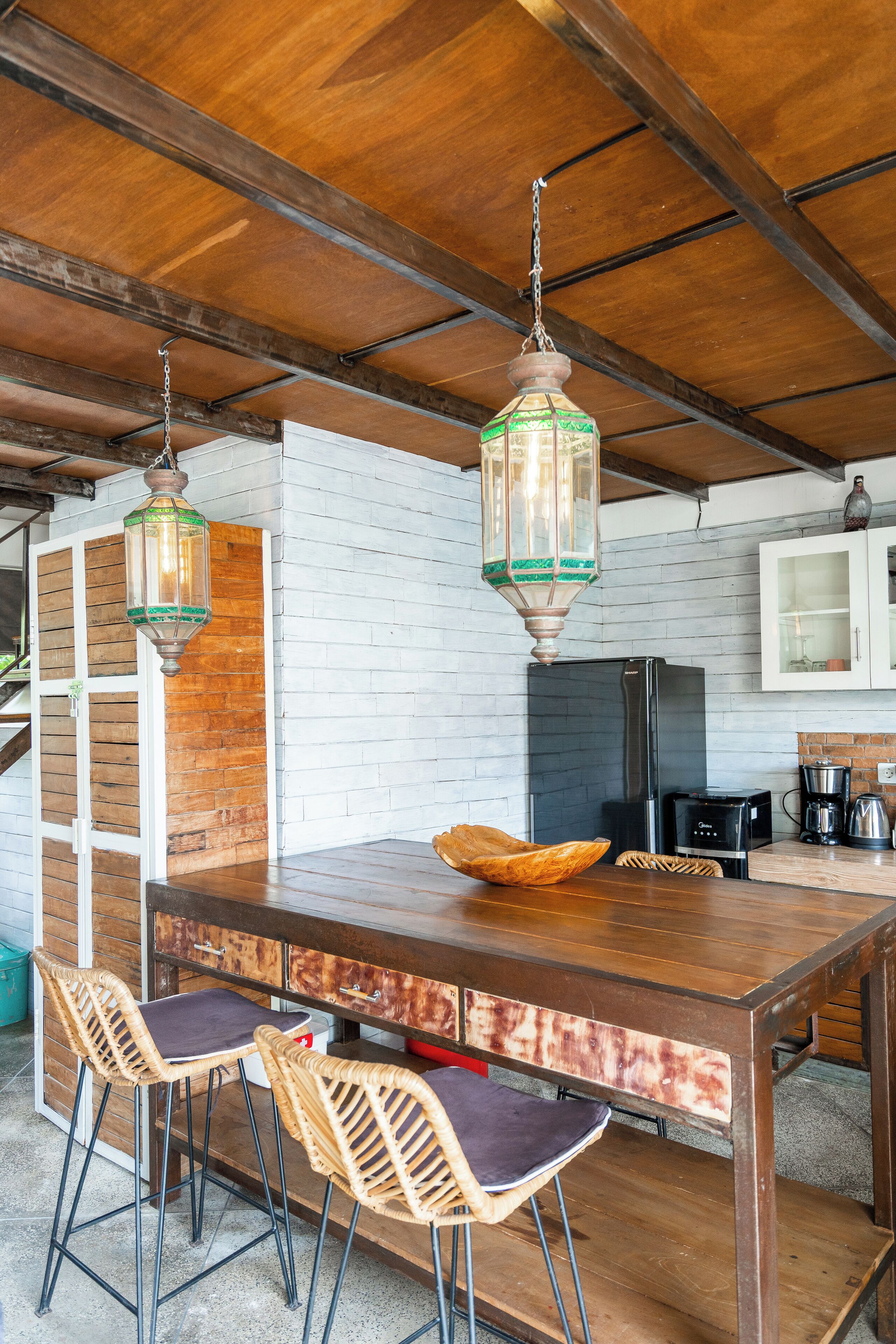A kitchen with a large island, two bar stools, hanging lanterns, and a refrigerator.