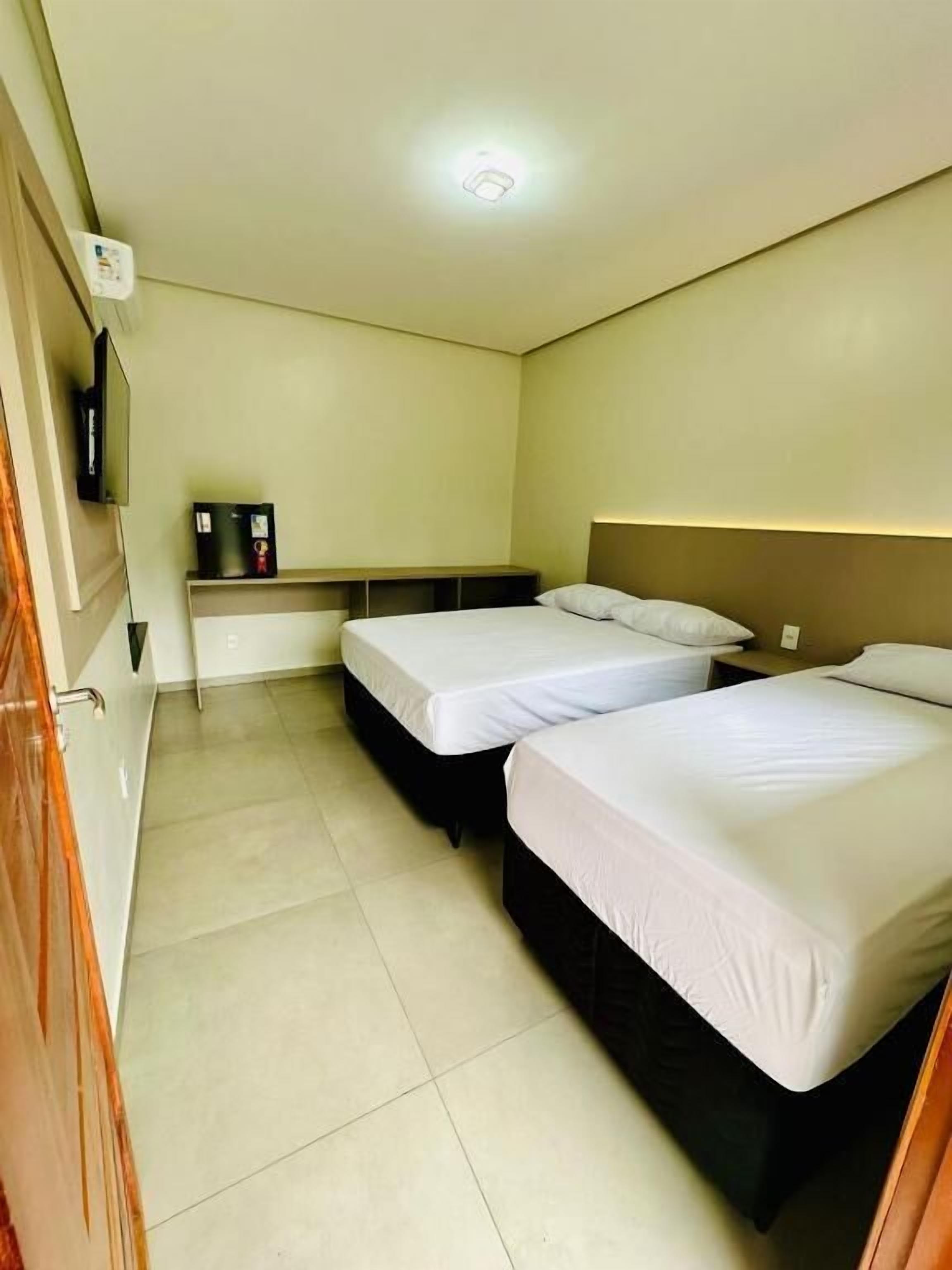 Habitación de hotel con dos camas, un televisor montado en la pared y un estante con objetos.