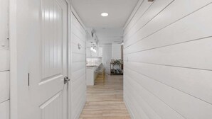 A narrow hallway with white paneling and a wooden floor, leading to a kitchen area with bar stools and a countertop.