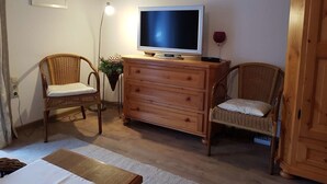Een woonkamer met een televisie op een houten kast, twee rieten stoelen, een bijzettafeltje en een aan de muur hangend schilderij.