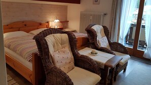 Een kamer met een bed, twee rieten stoelen, een tafeltje, een lamp en een raam met gordijnen.