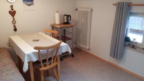 Een kleine kamer met een tafel, stoelen, een radiator, een raam met gordijnen, een klok en een aan de muur hangende foto.