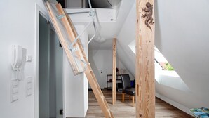 Ein Zimmer mit schräger Decke, Holzbalken, einer Leiter, einem Stuhl und einem Tisch mit einem Buch.