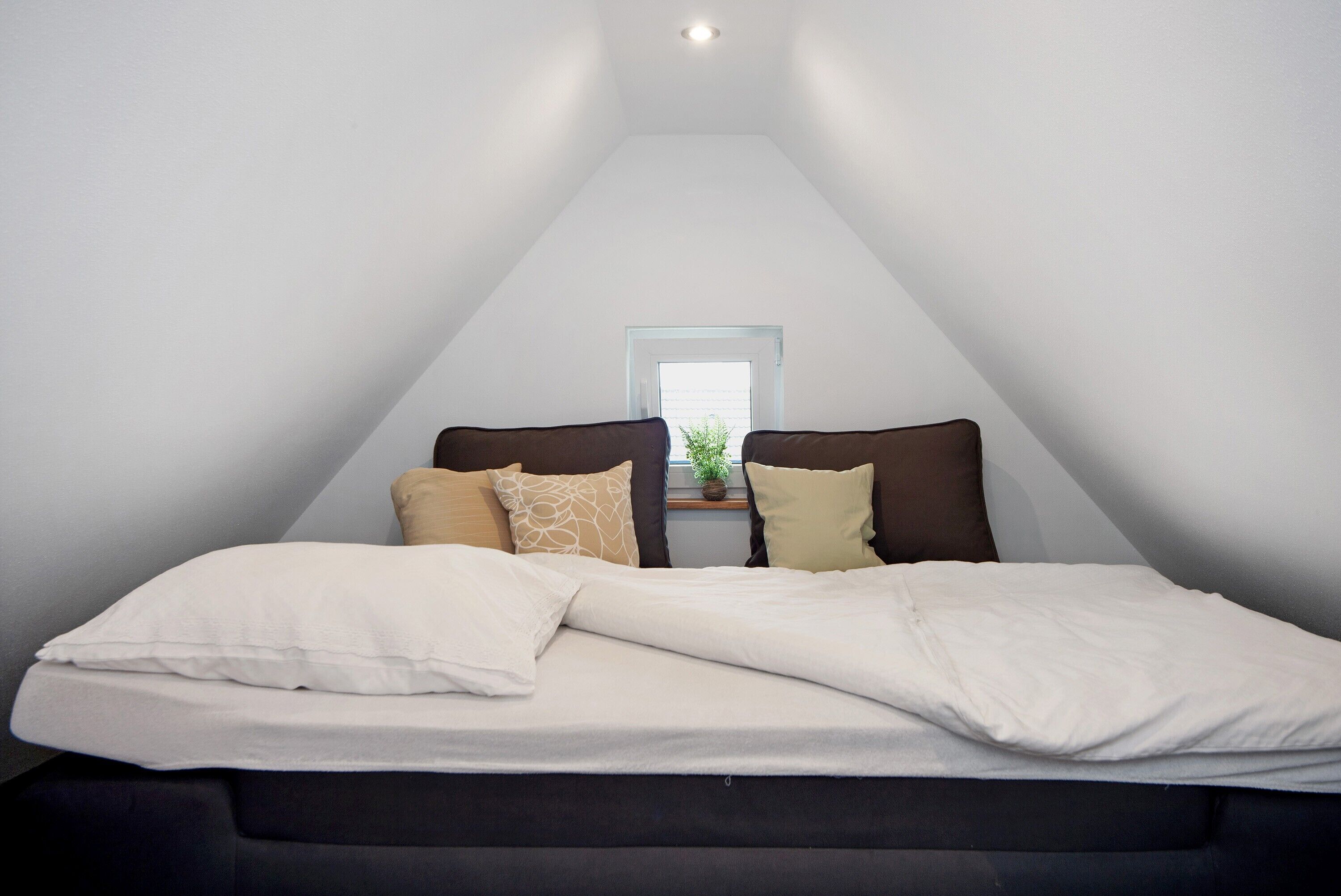 Une chambre avec un plafond en pente, un lit avec une literie blanche et une petite fenêtre donnant sur un paysage agréable.