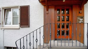 Ein Gebäudeeingang mit einer Holztür, einem Fenster mit Rollläden und einem Metallgeländer.