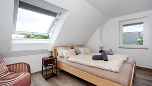 Ein Schlafzimmer mit schrägen Wänden, einem Bett mit Holzrahmen, einem Fenster, einem Sessel und einem Nachttisch.
