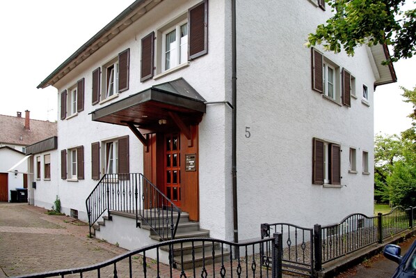 Ein zweistöckiges Haus mit Holztür, einer kleinen Veranda und der Hausnummer 5 an der Wand.