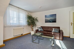 Una sala de estar con un sofá de cuero marrón, una mesa de centro de vidrio, una planta en maceta, un radiador y una ventana con cortinas traslúcidas.