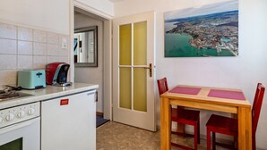Una cocina compacta con tostadora, cafetera y refrigerador. Hay una mesa de comedor con sillas y un cuadro de un paisaje urbano costero.